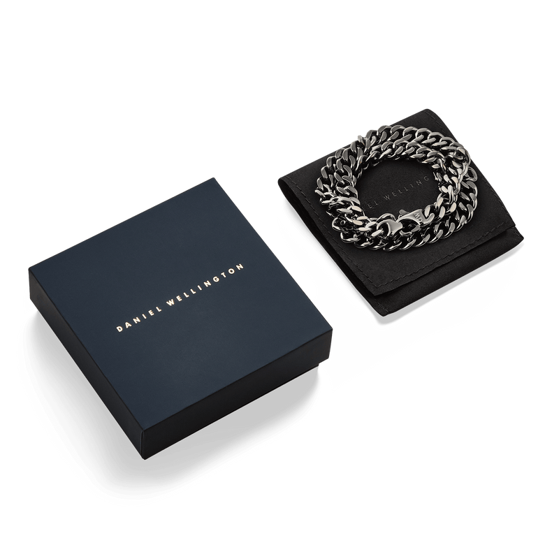 Ellipse Cuban Chain Necklace Graphite presented coiled on a black pouch next to a Daniel Wellington gift box, highlighting its bold links and matte graphite finish.