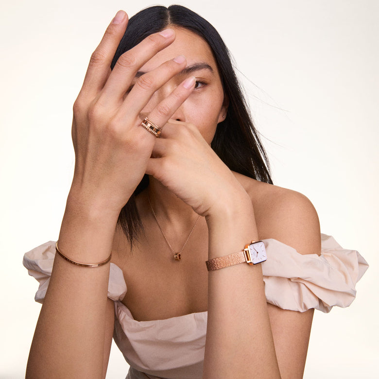 A person wearing the Elevation Ring Rose Gold, styled with a rose gold watch, bangle, and necklace, showcasing the ring’s bold geometric band and textured accents against a soft, off-the-shoulder top.