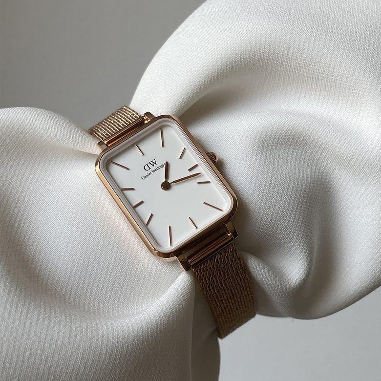 Quadro Pressed Melrose Rose Gold watch with a slim rectangular case, minimalist white dial, and rose gold mesh strap displayed elegantly on a textured white fabric background.
