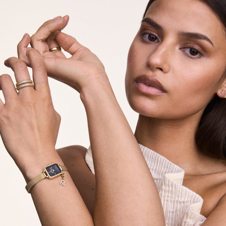 A woman wearing the Quadro Mini Crystal Zodiac Evergold gold watch with a black mother of pearl dial, crystal bezel, slim mesh strap, and zodiac charm, styled with gold rings and a cream textured top.