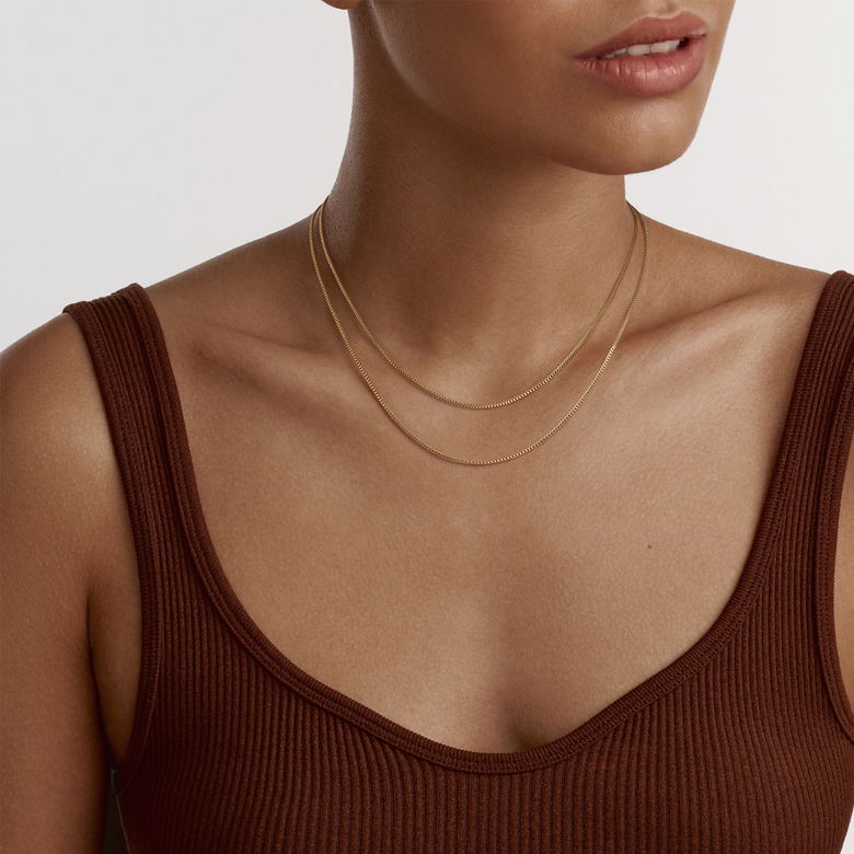 A woman wearing the Box Chain Necklace G, styled as a layered gold necklace with a slim, minimalist box chain design, complementing a brown ribbed top for a modern, elegant look.
