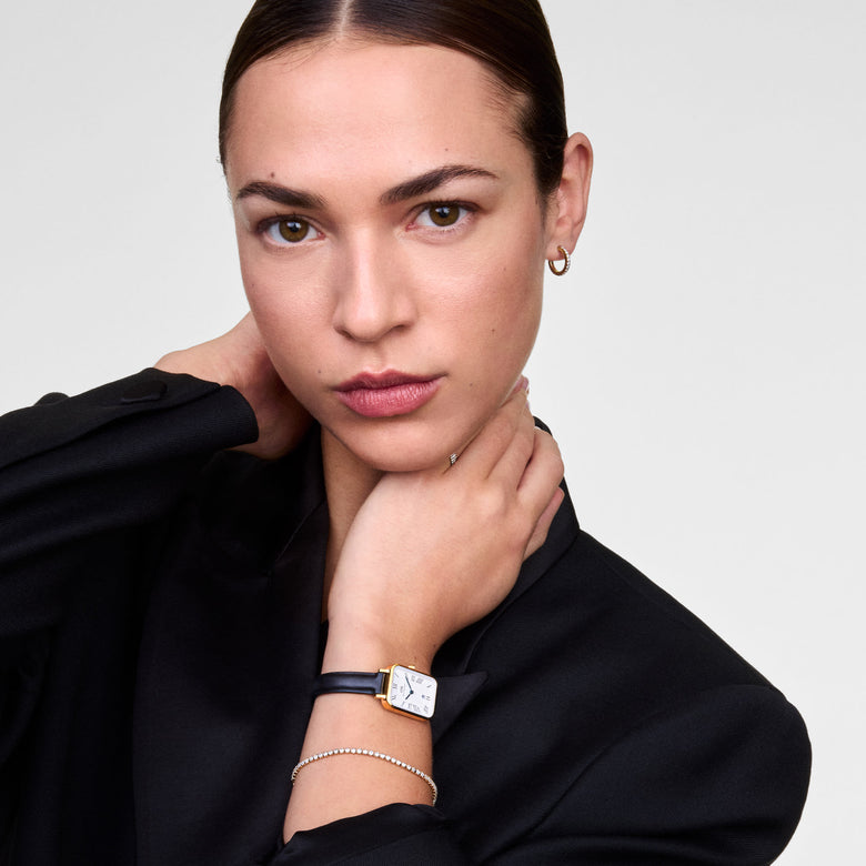 Quadro Roman Numerals Date Sheffield Gold ladies watch with a rectangular gold case and black leather strap, worn with a black blazer and a delicate bracelet for a refined, modern look.
