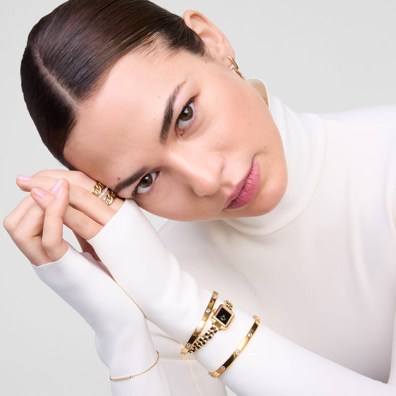 Quadro Mini Arch 3-link Onyx Sunray Gold ladies watch styled on a wrist with gold bracelets, highlighting its petite rectangular case, black dial, and polished gold bracelet against a white long-sleeve top.