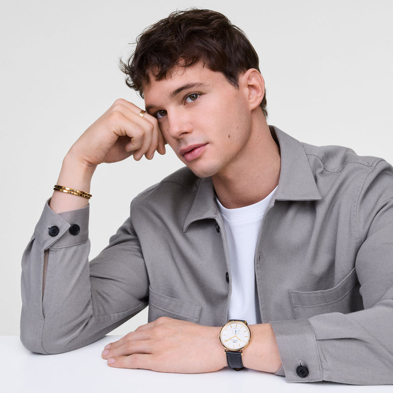 A man wearing the Classic Multi-Eye Sheffield Off-white Sunray Gold watch with a gold case, off-white dial, blue-tipped sub-second hand, and black leather strap, styled with a grey shirt and gold bracelet for a smart-casual look.
