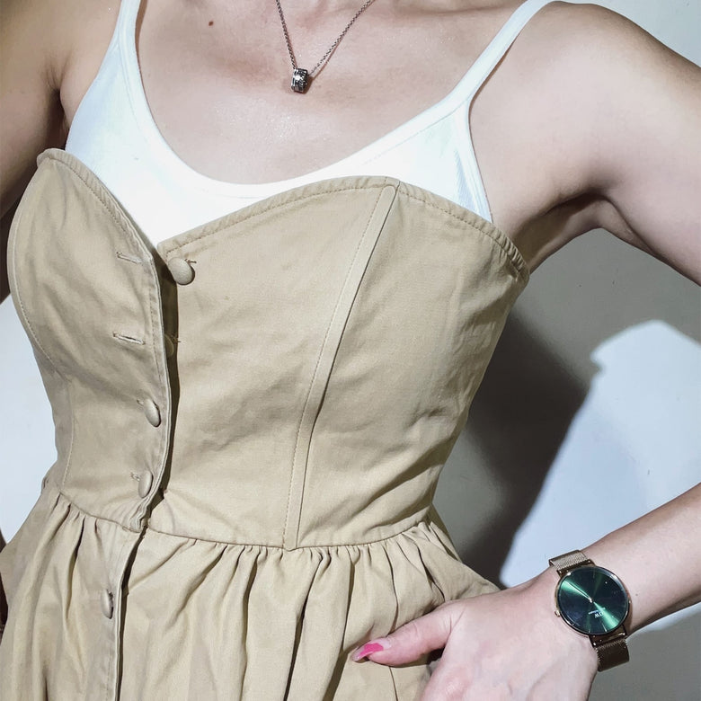 Woman wearing the Petite Emerald gold mesh strap watch with a deep green sunray dial, styled with a beige dress and white top for a modern, understated look.