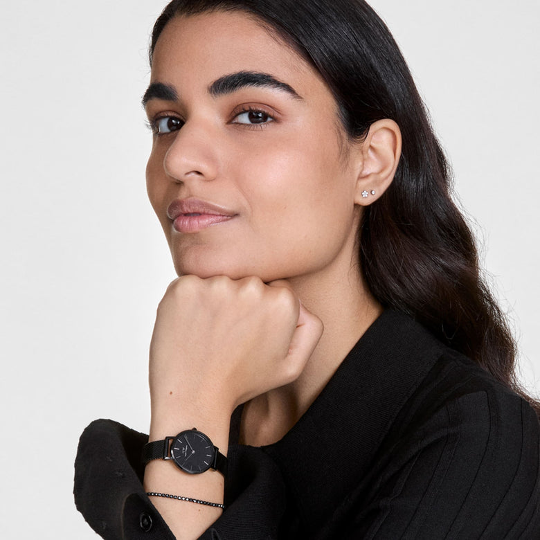 A woman wearing the Classic Tennis Bracelet Nightfall Rose Gold on her wrist, styled alongside a black watch and a black outfit, showcasing the slim rose gold bracelet with dark Preciosa crystals for a refined, minimalist look.