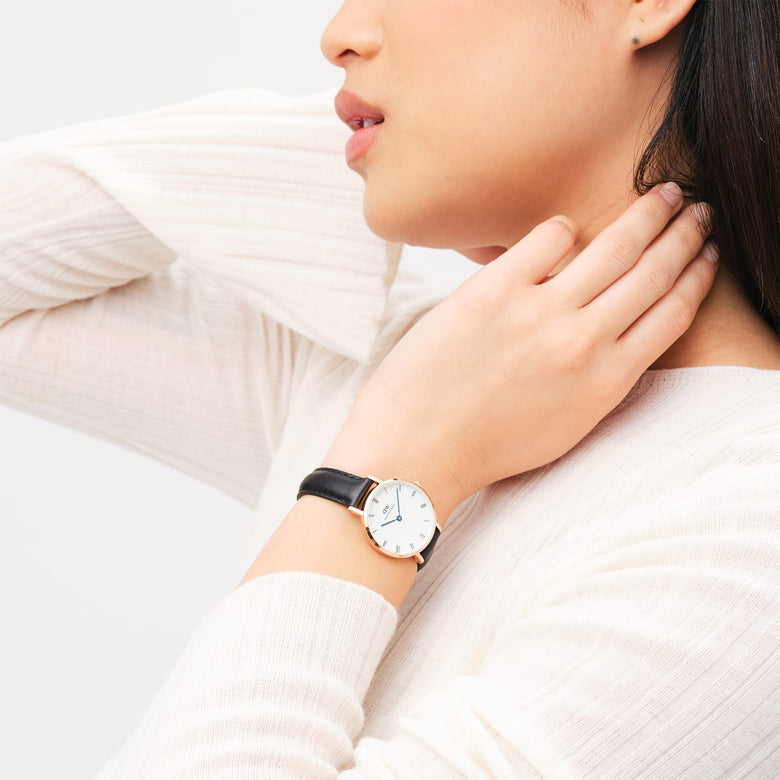 A woman wearing the Petite Roman Numerals Sheffield Rose Gold watch, showing its slim rose gold case, white dial with Roman numerals, blue steel hands, and black leather strap styled with a light long-sleeve top.