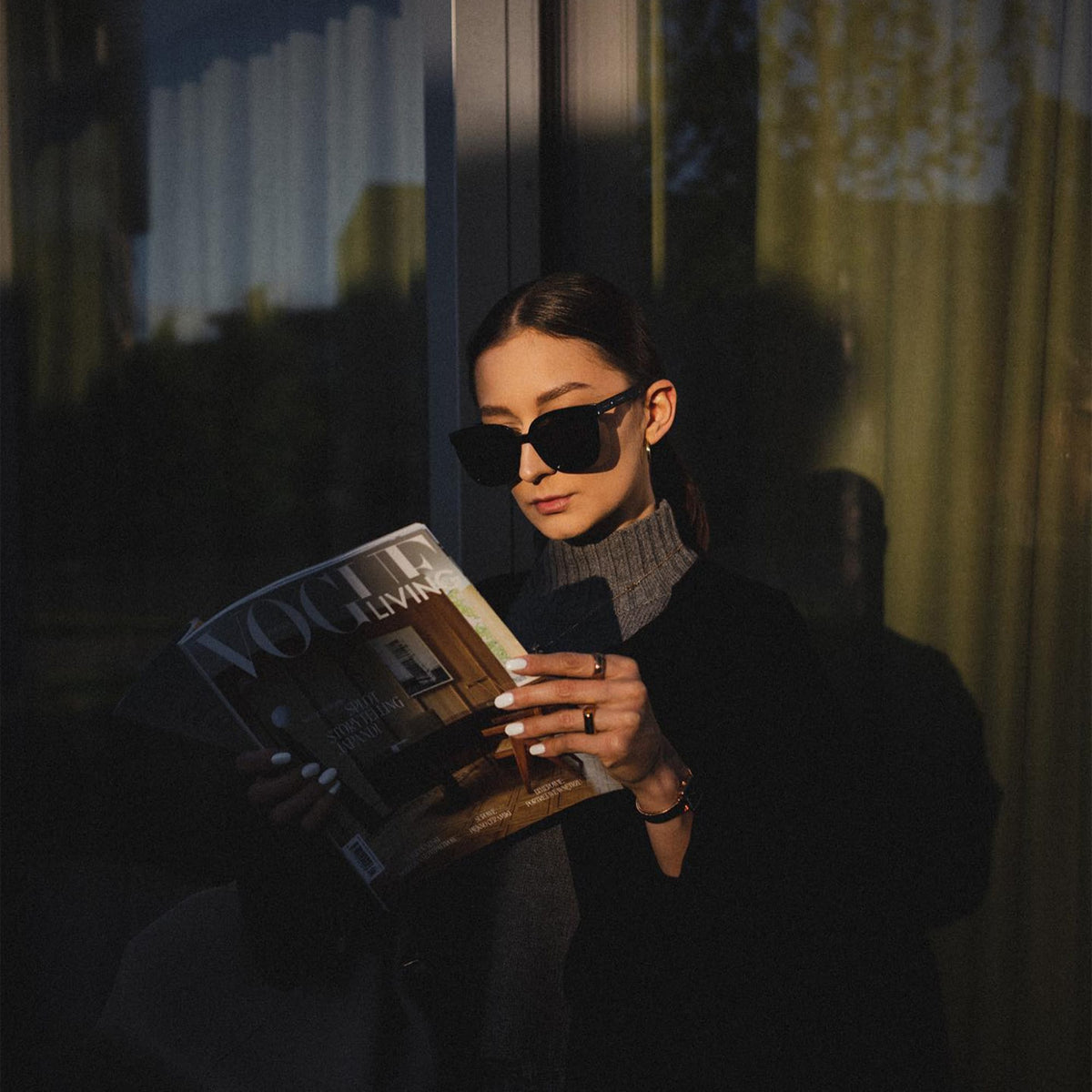 A person wearing Grande Bio-Acetate Black sunglasses with an oversized square frame and glossy finish, reading a magazine outdoors in natural light.