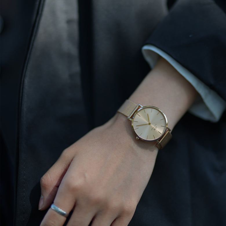 Petite Unitone Gold watch with a gold mesh strap and minimalist dial, worn on a small wrist and styled with a dark jacket for an elegant, everyday look.