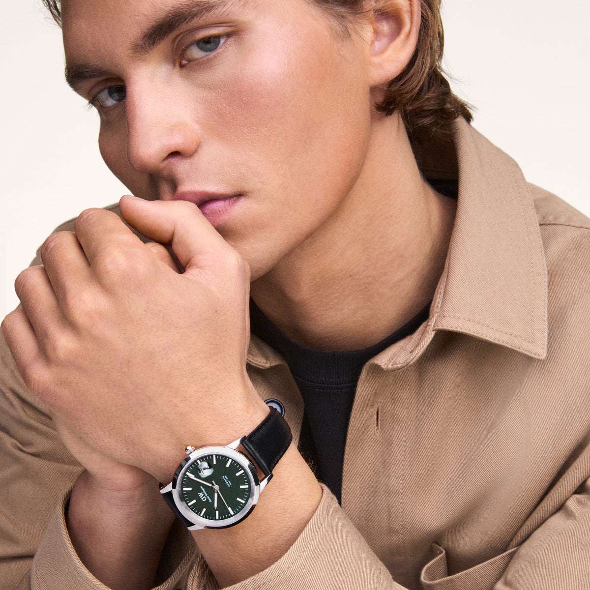 A man wearing the Daniel Wellington Iconic Sheffield Automatic Rose Gold watch, featuring a green enamel dial, rose gold case, and black leather strap, styled with a beige jacket for a smart-casual look.