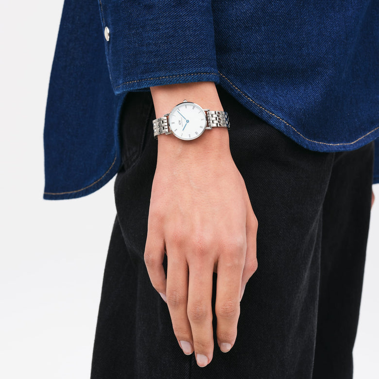 Petite Roman Numerals 5-Link Silver watch with a white dial, Roman numerals, and blue steel hands, worn on the wrist with a polished five-link stainless steel bracelet, styled casually with a denim shirt and black trousers.