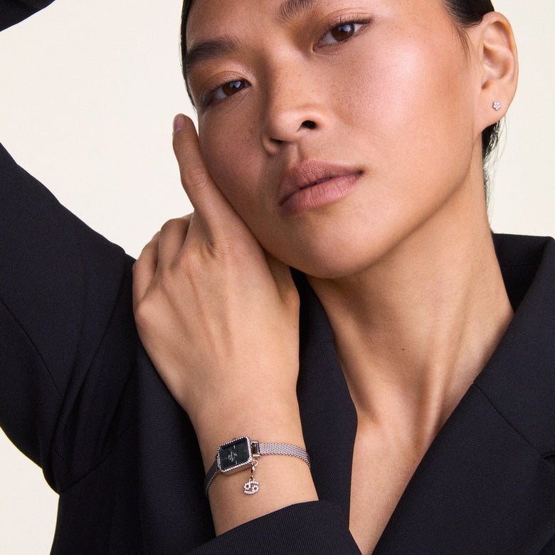A woman wearing the Quadro Mini Crystal Zodiac Sterling Set, featuring a silver mesh strap, rectangular crystal-framed case, black mother of pearl dial, and a detachable zodiac charm, styled with a black blazer for a modern, elegant look.