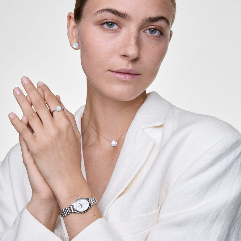A woman wearing Daniel Wellington Audrey Earrings Mother of Pearl Silver, styled with a matching necklace, ring, and silver watch, showcasing the elegant mother of pearl centre and sparkling cubic zirconia halo in a polished, modern look.