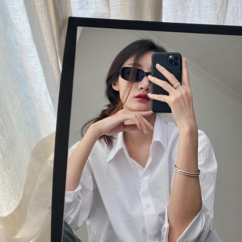 A woman wearing a white shirt takes a mirror selfie, showcasing the Daniel Wellington Emalie Ring Satin White Rose Gold on her finger, styled with a matching rose gold bangle for a modern minimalist look.