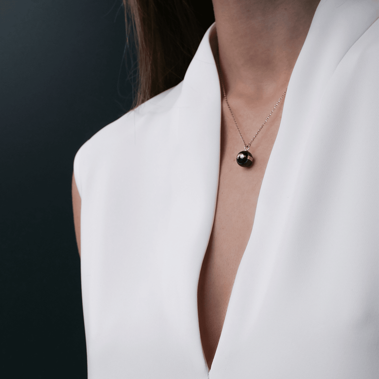 A woman wearing the Aspiration Necklace Rose Gold Black, featuring a polished black ceramic pendant with a rose gold plated band, styled with a white V-neck blouse for a modern and elegant look.