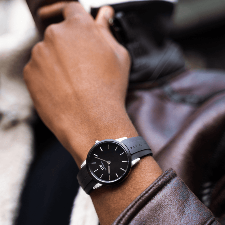 A person wearing the Daniel Wellington Iconic Motion Silver watch with a black dial and black FKM rubber strap, styled with a brown leather jacket for a modern, everyday look.