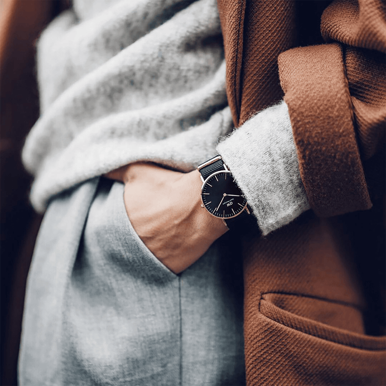 A person wearing the Classic Cornwall Black Rose Gold watch, styled with a grey jumper, light trousers, and a brown coat, highlighting the watch’s black NATO strap and rose gold case for a refined everyday look.