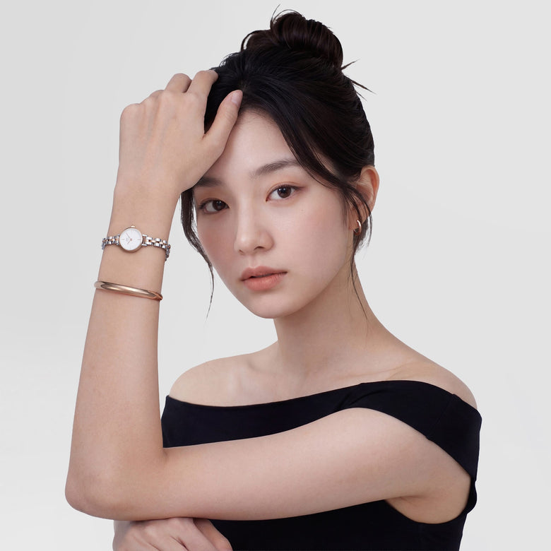 A woman wearing the Petite Mini Arch 3-Link Two Tone Rose Gold watch, styled with a rose gold bangle and an off-the-shoulder black top, showcasing the elegant two tone bracelet and minimalist white dial.