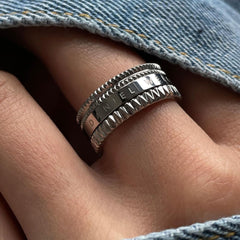 The Elevation Ring Silver by Daniel Wellington is shown worn on a finger, featuring a wide polished silver band with geometric textured edges and an engraved centre, styled with casual denim for a modern look.
