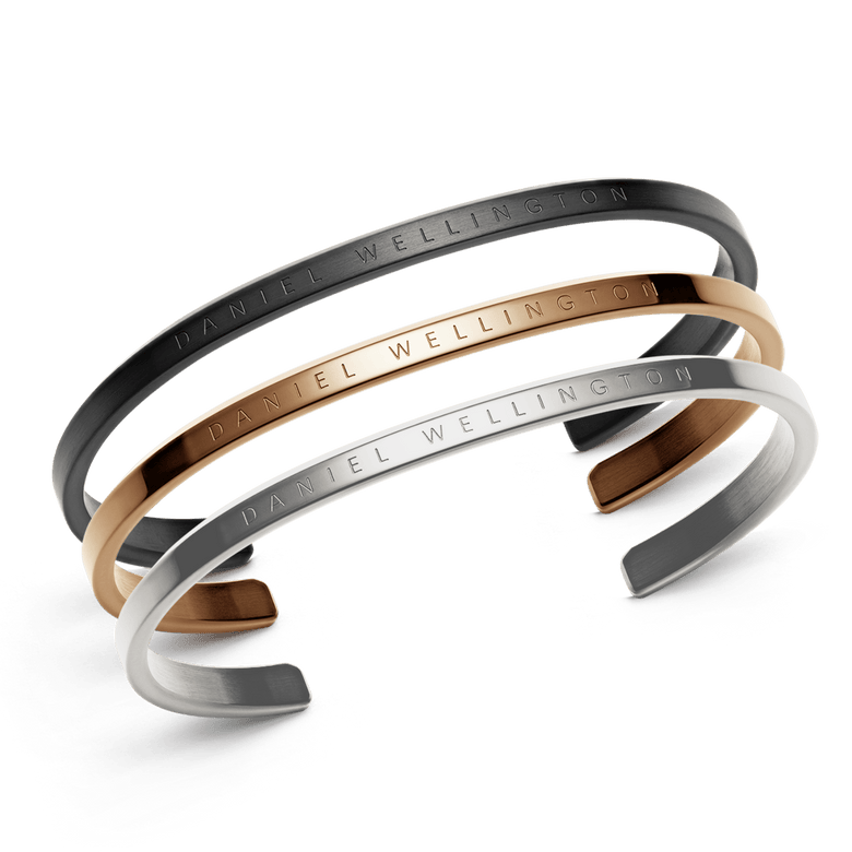 Three minimalist cuff bracelets in black, rose gold, and silver stainless steel, each engraved with Daniel Wellington branding and featuring a sleek, adjustable design suitable for layering or wearing alone.