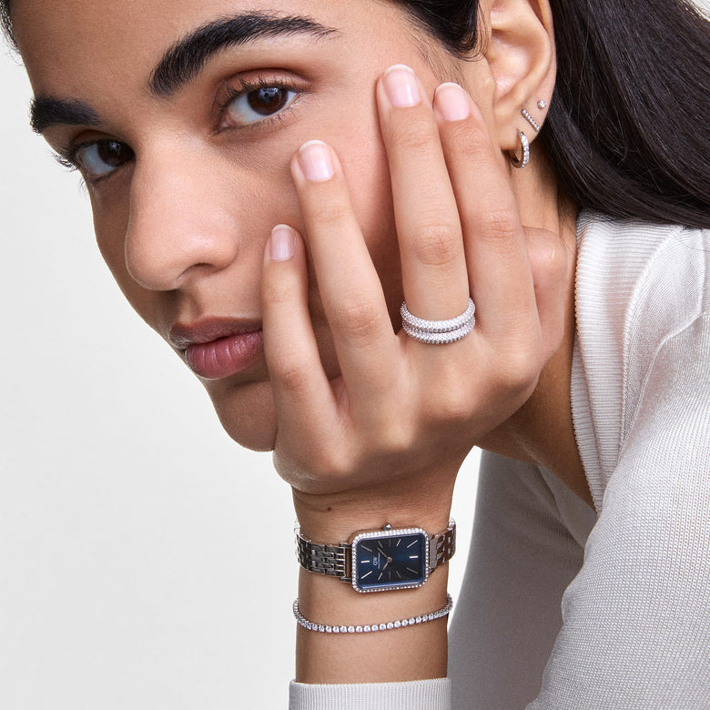 A woman wearing the Classic Tennis Bracelet Silver on her wrist, styled with a silver watch and matching jewellery, showcasing the bracelet’s slim design and sparkling cubic zirconia crystals in a modern, elegant look.