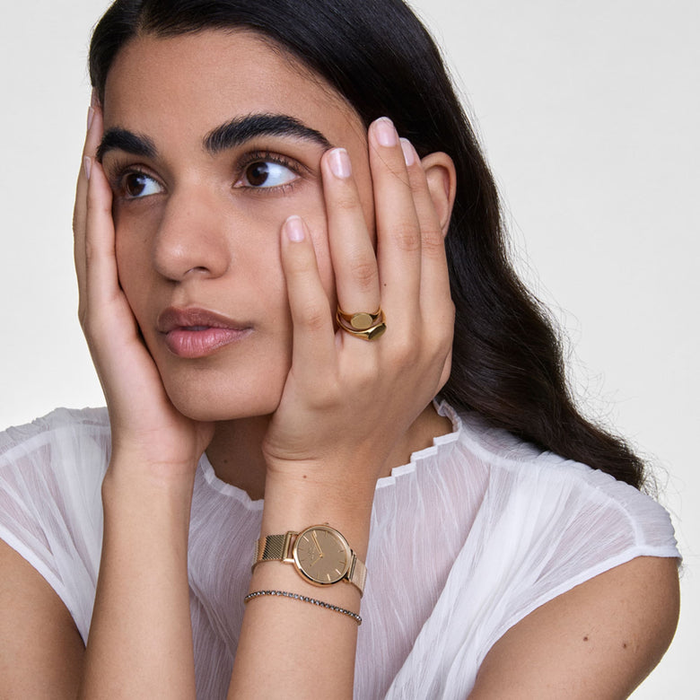 A woman wearing the Daniel Wellington Petite Reflection Gold watch with a gold mesh strap and mirror-effect dial, styled alongside a delicate bracelet and a sheer white blouse for an elegant everyday look.