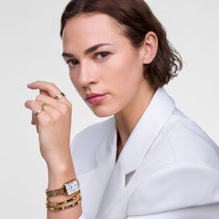 A woman wearing a white blazer showcases the Quadro Roman Numerals Date gold watch with a rectangular face and Roman numeral dial, styled alongside multiple Classic Lumine gold bracelets with crystal details for an elegant, layered look.