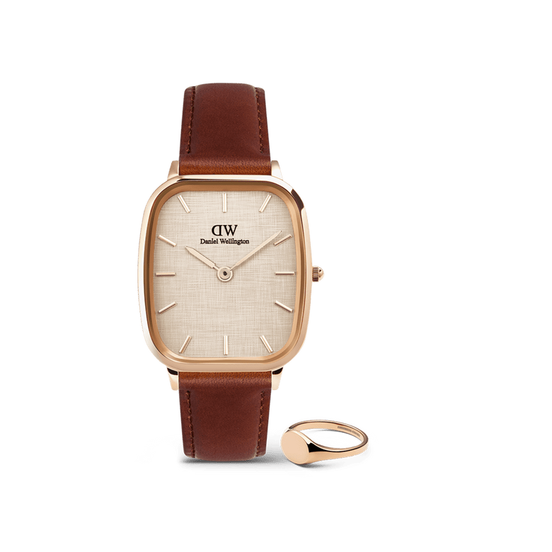 Rose gold Daniel Wellington watch with a cushion-shaped case, textured yellow dial, and brown leather strap, paired with a polished rose gold mini signet ring, both displayed against a plain background.