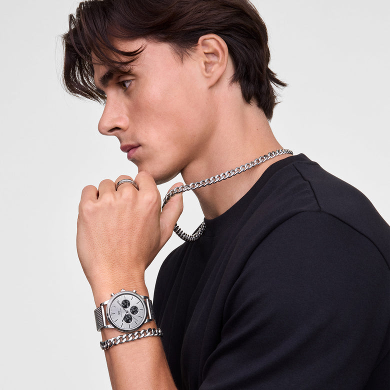 A man wearing the Daniel Wellington Iconic Chronograph Panda watch with a silver mesh strap and silver subdials, paired with the Ellipse Cuban Chain Bracelet and matching chain necklace, styled with a black t-shirt for a modern, coordinated look.