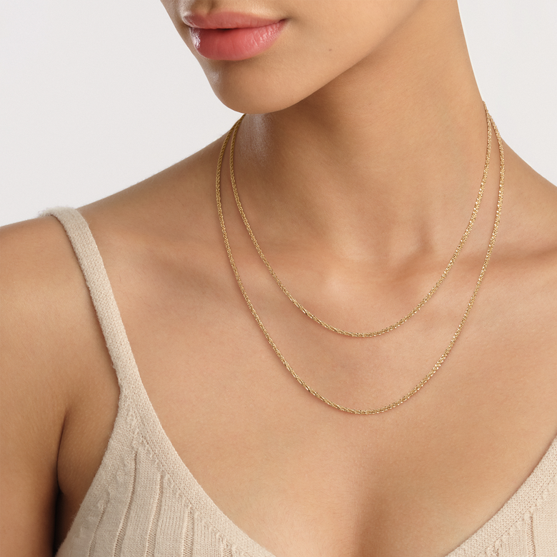 Twisted Chain Necklace G with a gold finish, styled as a layered look on a woman wearing a beige top, highlighting its slim, textured design and versatility for everyday wear.