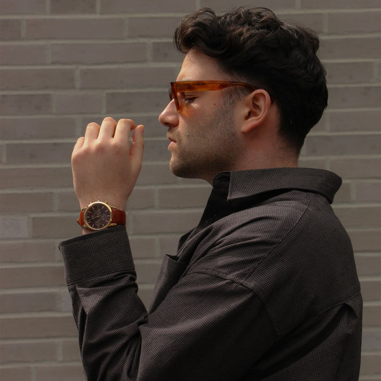 A man wearing the Iconic Chronograph St Mawes Amber RG watch with a rose gold case, amber dial, and brown leather strap, styled with a dark shirt in a casual outdoor setting.