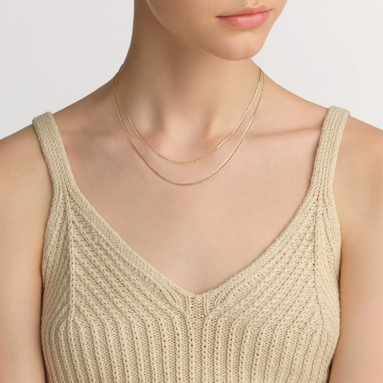 Flat Chain Necklace G in gold, styled as a layered necklace on a woman wearing a beige knitted top, showcasing its slim, minimalist profile and elegant finish.