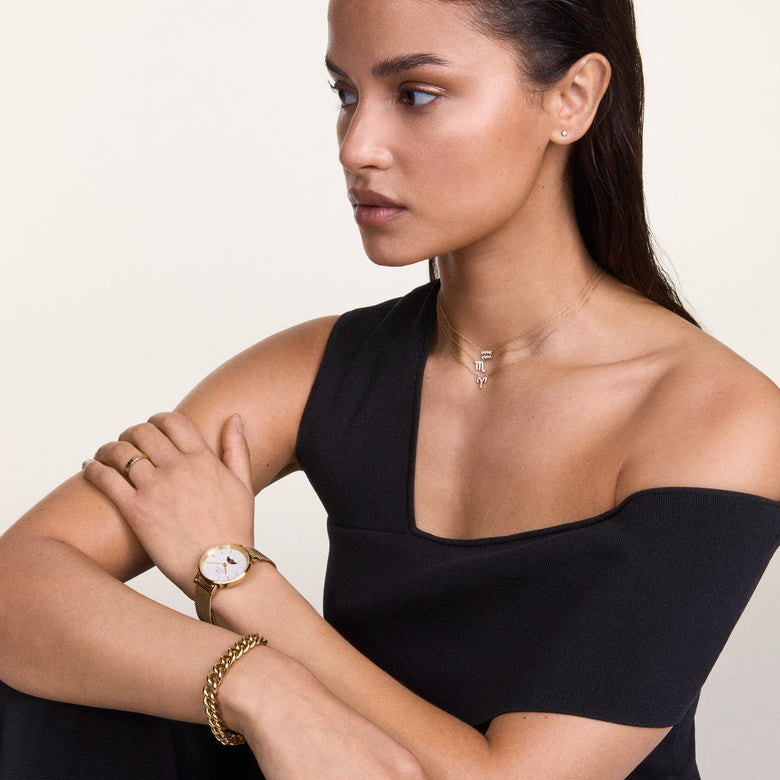 A woman wears the Crystal Zodiac Chain Necklace Silver with a Scorpio zodiac pendant, styled with layered necklaces, a gold watch, and a black off-shoulder top for a modern, elegant look.