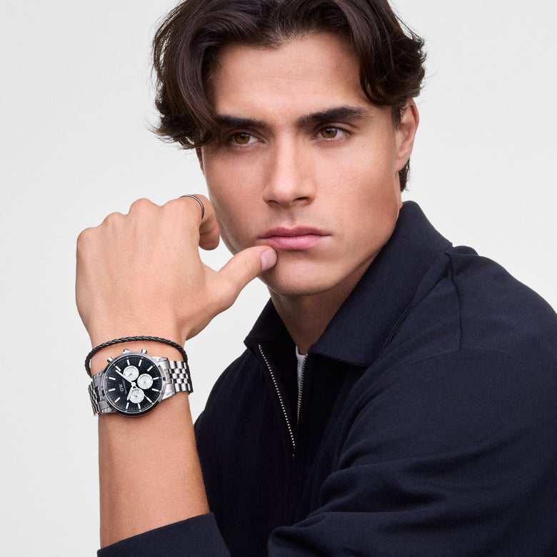 A man wearing the Iconic Chronograph Arch 5-Link Onyx Panda Silver watch with a polished stainless steel bracelet and black dial with white sub-dials, styled alongside a braided bracelet and a navy zip-up top.