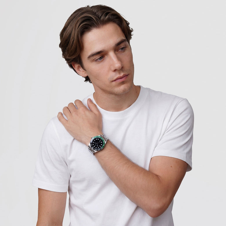 A man wearing the Daniel Wellington Iconic GMT Link Black Green Ceramic Bezel Silver watch, styled with a casual white t-shirt, showing the black dial, green and black ceramic bezel, and silver stainless steel bracelet on his wrist.