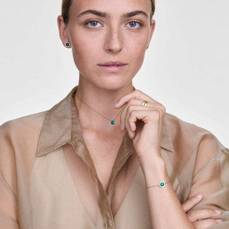A woman wears the Audrey Bracelet Malachite Gold on her wrist, showcasing its slim gold chain and green malachite centrepiece with a halo of cubic zirconia, styled with a sheer beige blouse and matching malachite jewellery for a coordinated, elegant look.