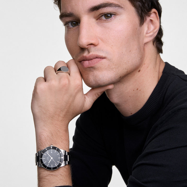 A man wearing the Daniel Wellington Iconic Paradigma Black & Classic Signet Ring Set, featuring a polished silver signet ring with a black face and a stainless steel watch with a black dial, styled with a black jumper for a modern, refined look.