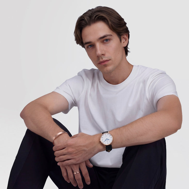 A man wearing the Classic Roman Numerals Date Black Croc Belly Rose Gold watch, styled with a white t-shirt and black trousers, showing the rose gold case, white dial with Roman numerals, blue hands, and black croc-embossed leather strap.