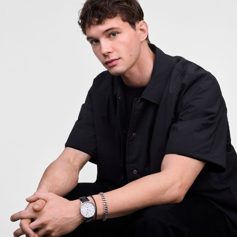 A man wearing the Classic Multi-Eye Sheffield Off-white Sunray Silver watch with a black leather strap, styled with a silver bracelet and short-sleeve black shirt, highlighting the watch’s minimalist dial and blue-tinted sub-second hand for a modern, versatile look.