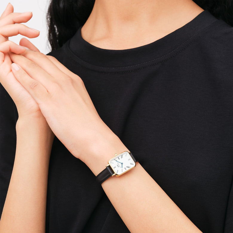 Quadro Roman Numerals Sheffield Gold ladies watch with a rectangular gold case, white dial, Roman numerals, and slim black leather strap, worn on the wrist with a casual black top.