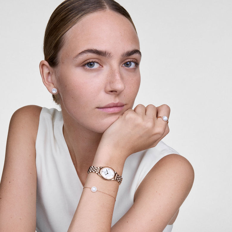 A woman wearing the Audrey Mother Of Pearl Gold Set, including a gold bracelet, ring, and stud earrings each featuring a mother of pearl centre with a halo of sparkling stones, styled with a sleeveless white top and a gold watch for an elegant, coordinated look.
