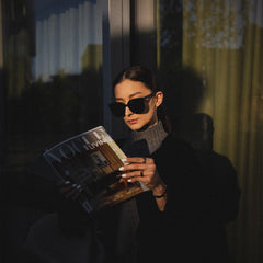A person wearing Grande Bio-Acetate Black sunglasses with an oversized square frame and glossy finish, reading a magazine outdoors in natural light.