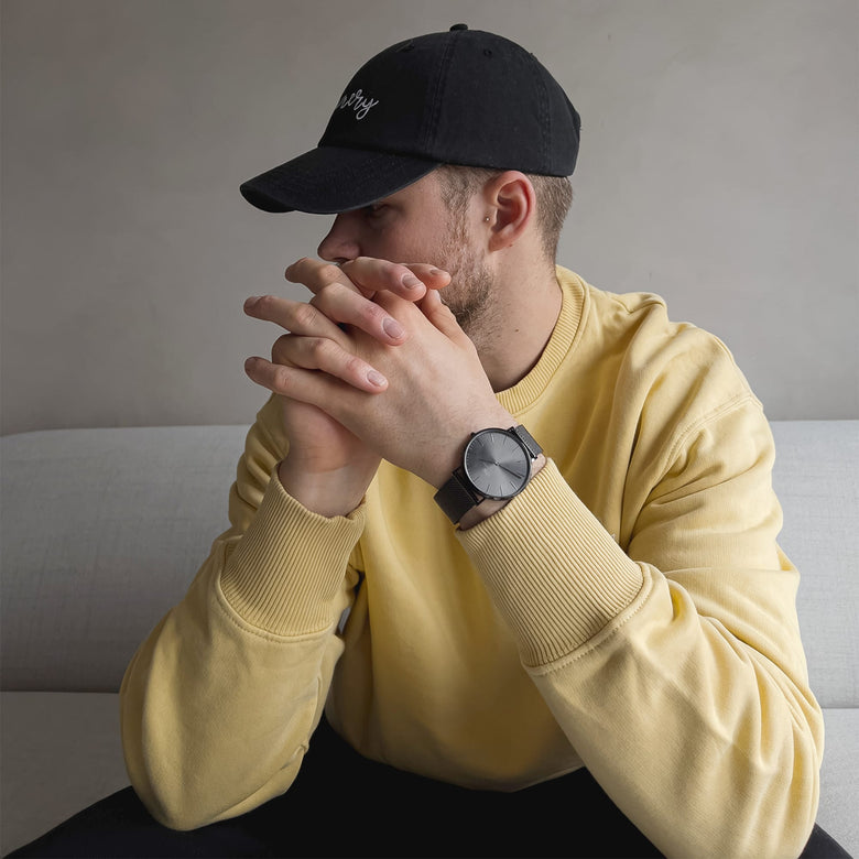 A man wearing a yellow jumper and black cap is seated on a sofa, showcasing the Classic Mesh Graphite watch with its graphite grey stainless steel mesh strap and minimalist sunray dial on his wrist.