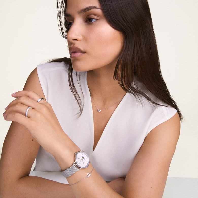 A woman wearing the Crystal Zodiac Chain Necklace Silver, featuring a delicate silver chain and a sparkling zodiac pendant, styled with a white sleeveless top and complemented by matching silver jewellery.
