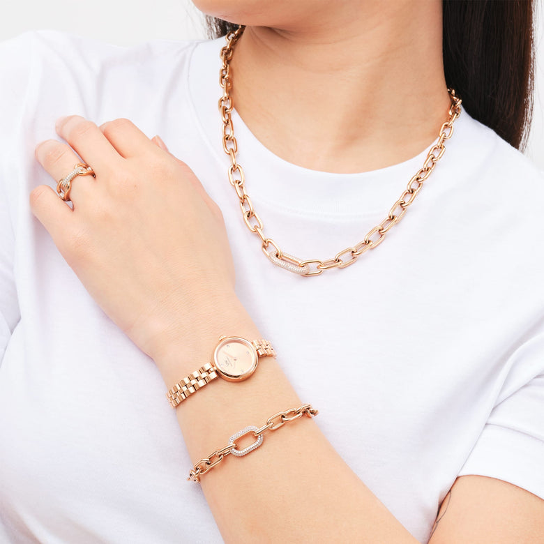 A woman wearing the Crystal Link Ring Rose Gold on her finger, styled with a matching rose gold chain necklace, bracelet, and watch, all set against a white t-shirt for a modern, coordinated look.