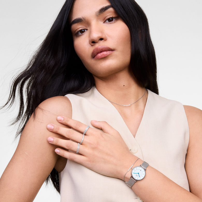 A woman wears the Petite Roman Numerals Date Silver watch with a slim silver mesh strap, blue hands, and Roman numerals, styled with minimalist jewellery and a neutral sleeveless top for a modern, elegant look.