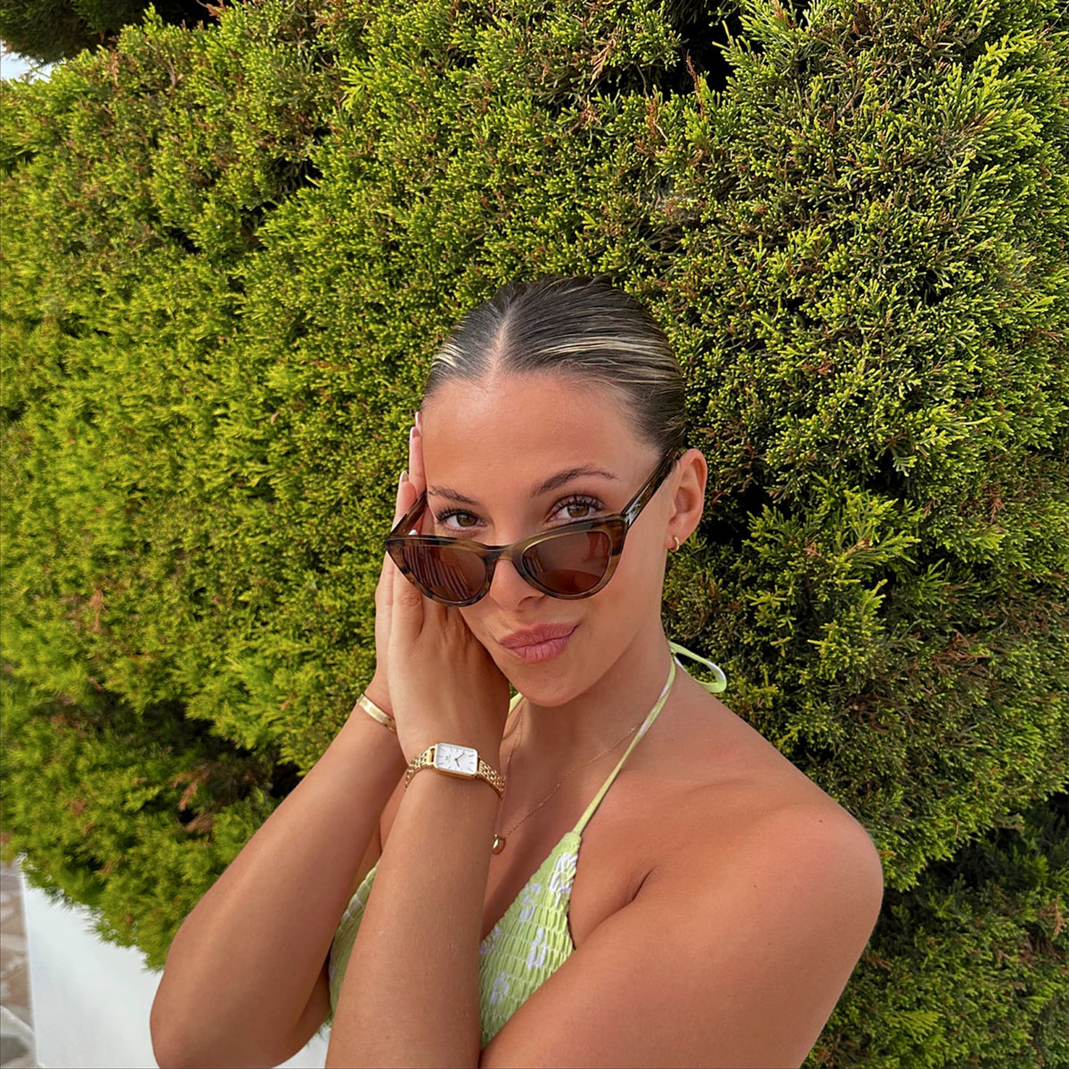 A woman wearing Lynx Bio-Acetate Brown Demi sunglasses with a cat eye frame and amber lenses, styled outdoors in a green patterned top and accessorised with a gold watch and bracelet.