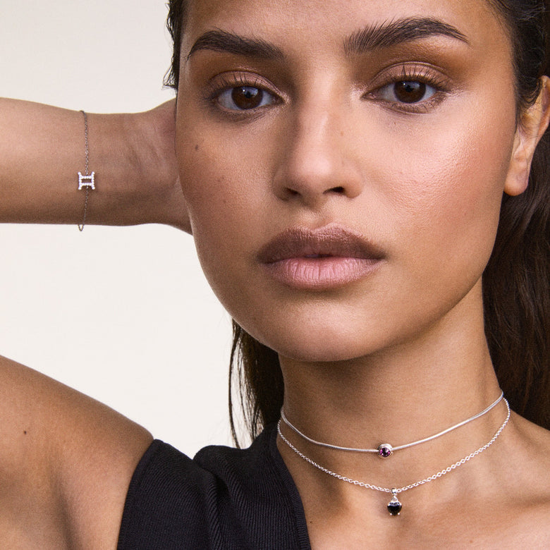 A woman wears the Crystal Zodiac Chain Bracelet Silver on her wrist, showcasing its delicate silver chain and crystal-encrusted Gemini zodiac charm, styled with layered silver necklaces for a modern, minimalist look.