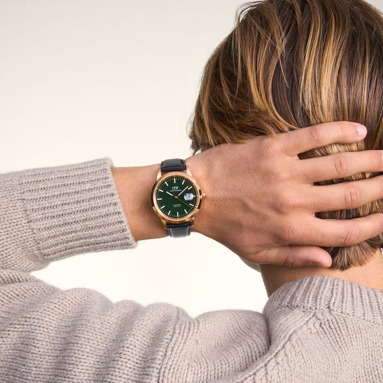 A person wearing the Daniel Wellington Iconic Sheffield Automatic Rose Gold watch with a green enamel dial and black leather strap, styled with a light beige knitted jumper for a relaxed, modern look.