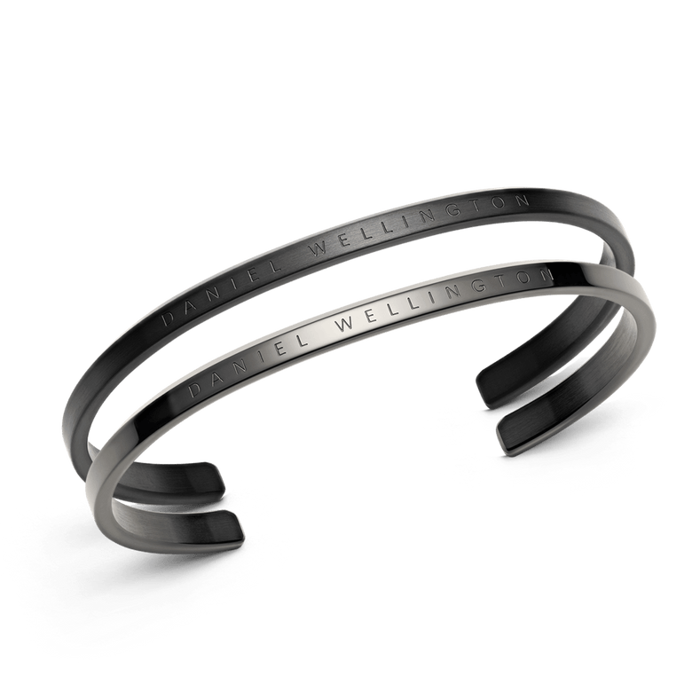 Classic Bracelet Black + Graphite set featuring two minimalist stainless steel cuff bracelets in matte black and graphite finishes, each engraved with Daniel Wellington branding.